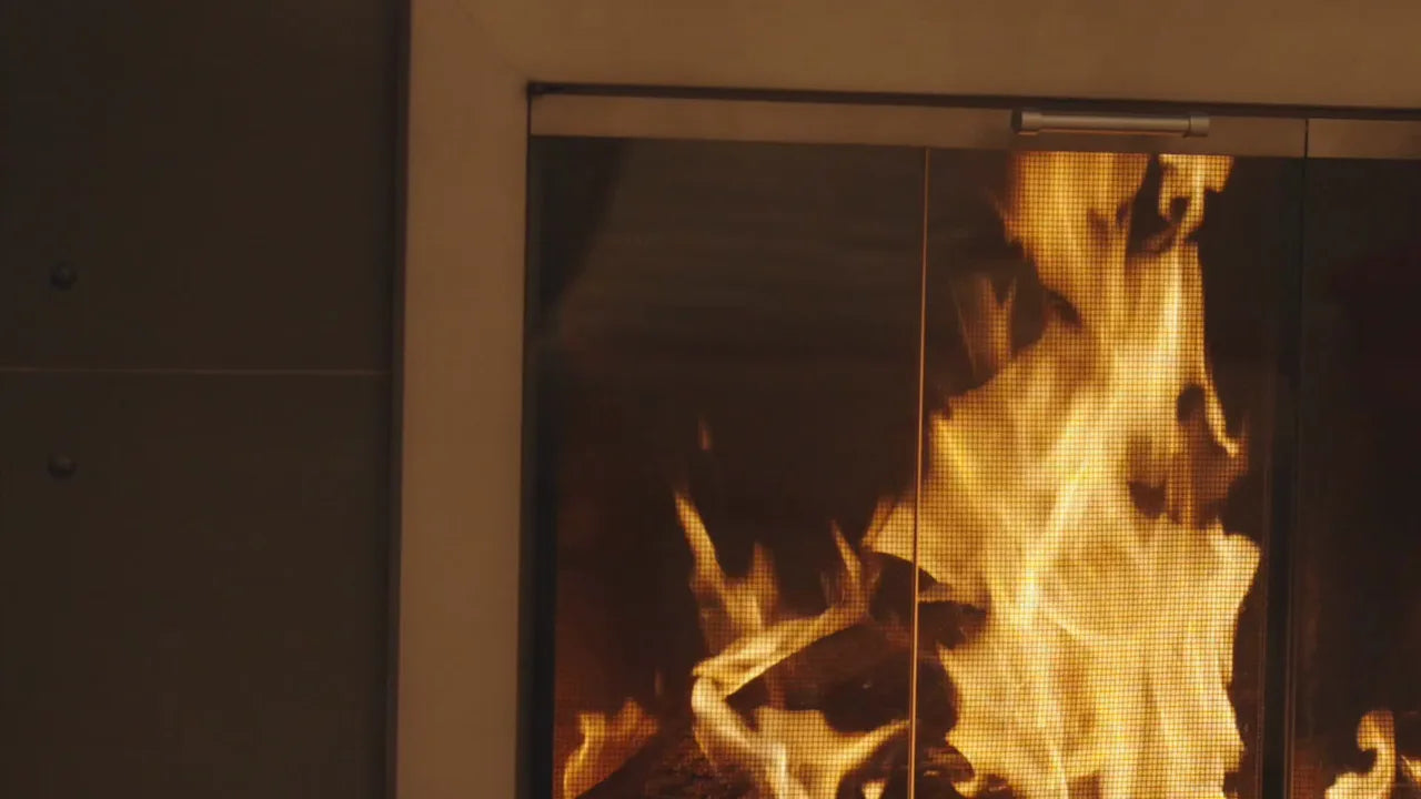 A lit fire behind bronze bifold fireplace doors, in a furnished room.