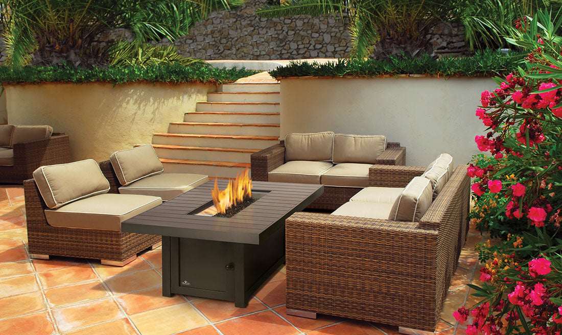 Napoleon St. Tropez rectangular fire table burning beside wicker seating on a tiled patio