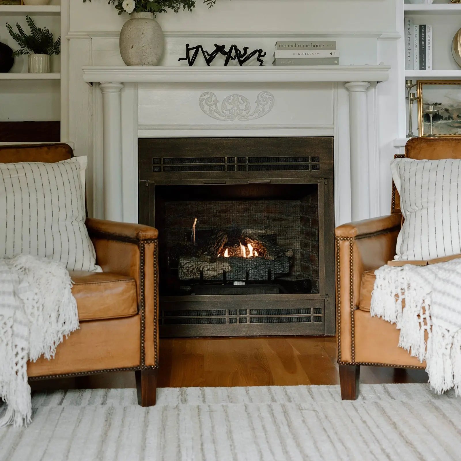 A gas log set burning behind a bronze fireplace door, between two leather club chairs